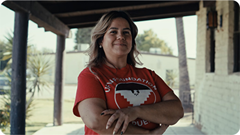 A confident woman standing with arms crossed wearing a red shirt with a UFW Foundation logo, in a relaxed outdoor setting with a blurred background.