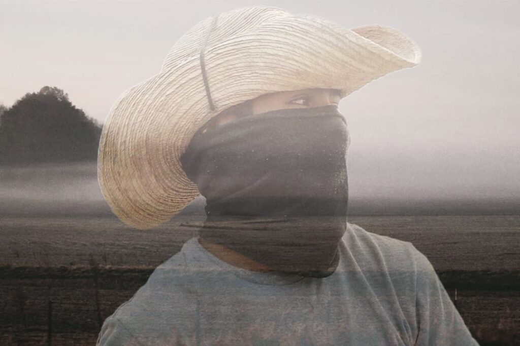 A man wearing a large straw hat and a face covering standing in a misty field.